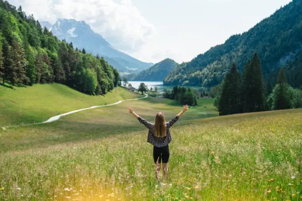 Frau steht mit erhobenen Armen in einem Feld mit Bergen im Hintergrund.