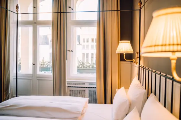HENRI-Berlin-Boutique-Hotel-Zimmer-Design Gemütliches Hotelzimmer im HENRI Berlin mit hohen Decken, hellem Fenster und eleganten Nachttischlampen im Gründerzeit-Stil.