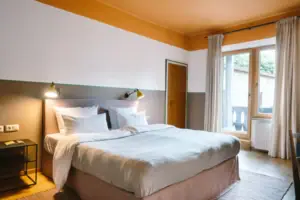 Ein gemütliches Bett in einem modernen Hotelzimmer im HENRI Hotel Garmisch-Partenkirchen mit einem Zugang zum Balkon im Hintergrund.