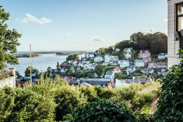 Stadtansicht mit Wasserfläche im Vordergrund, umgeben von Gebäuden und Bäumen unter einem bewölkten Himmel.